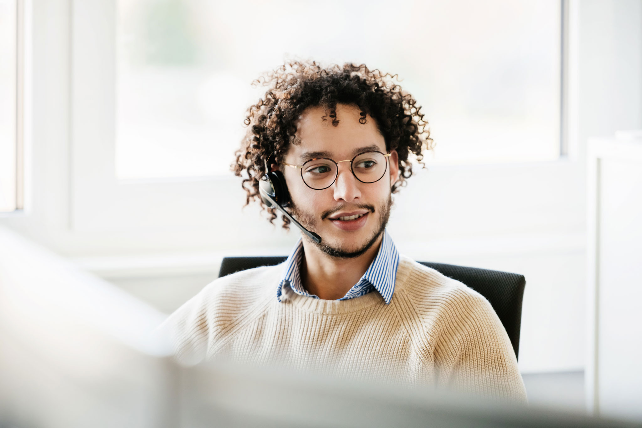 A male office employee wearing a wireless headset used to talk to clients, working at his computer in the office.